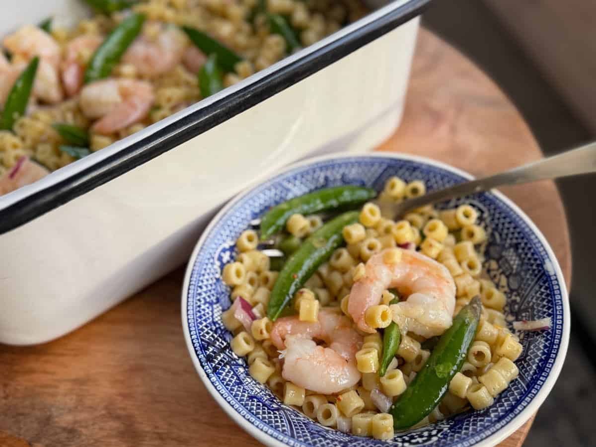 A bowl of pasta salad with shrimp, snap peas, and chopped onions is placed next to a larger white serving dish with similar contents. A fork is in the bowl, and the setup is on a wooden surface.