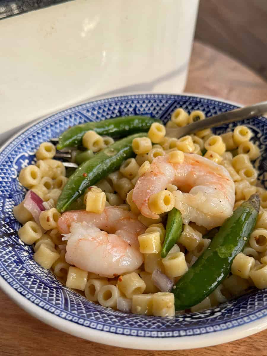 A bowl of pasta salad featuring small tube-shaped pasta, cooked shrimp, and sugar snap peas. The salad is served in a blue and white patterned bowl, placed on a wooden surface.