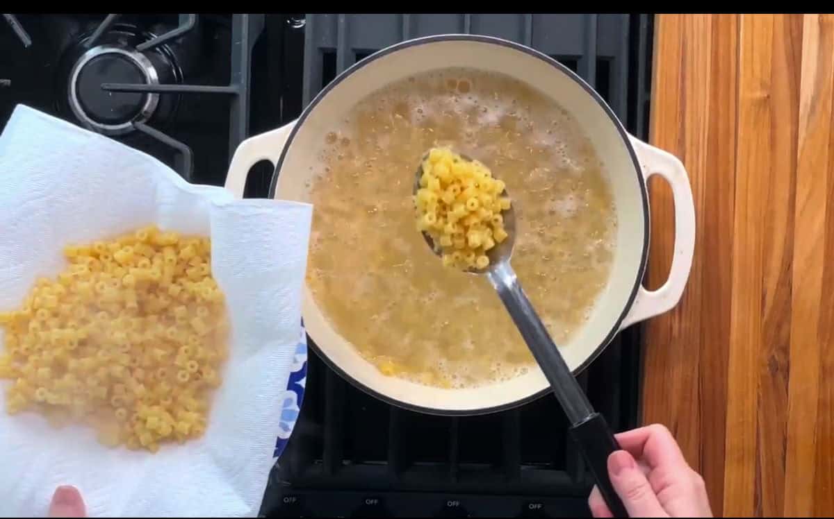 A hand holds a slotted spoon lifting cooked macaroni pasta from a boiling pot on a stove. Another hand holds a paper towel with drained macaroni on a plate. The pot is white and the stove burners are visible.