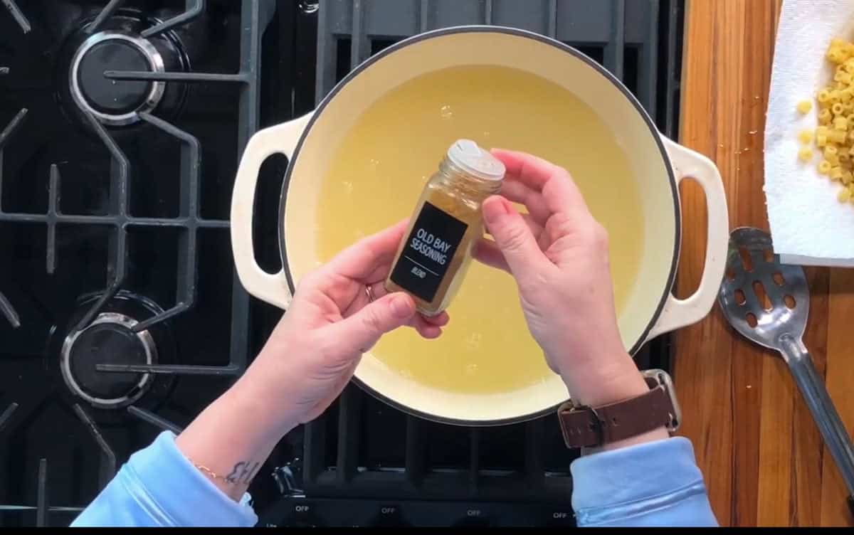 Hands holding a jar of "Old Bay" seasoning over a white pot filled with liquid on a stove. A slotted spoon and small pasta pieces on a paper towel are on the wooden counter to the right.