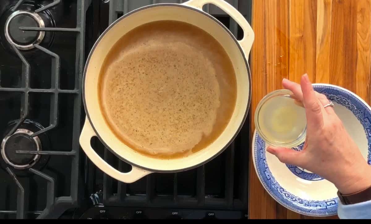 A person holds a small bowl of liquid over a pot of brown broth on a stove. The stove is black with three visible burners, and the pot is white with two handles. A wooden countertop with a blue and white plate is adjacent to the stove.