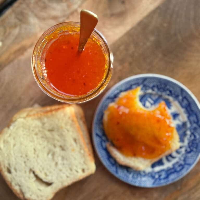 A jar of bright orange jam with a spoon placed inside is on a wooden surface. Next to it is a slice of bread and a blue and white plate with a piece of bread spread with the same jam.