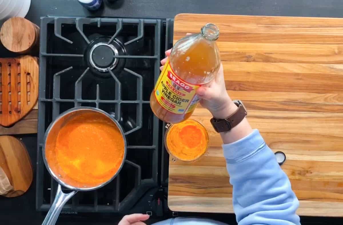 A person is holding a bottle of apple cider vinegar above a glass on a wooden countertop. A saucepan with an orange liquid is on a stove. The countertop also has a wooden board and a blue surface is visible.