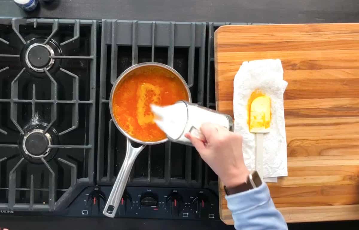 A person is pouring milk from a metal pitcher into a saucepan containing an orange-colored mixture. The saucepan is on a gas stove. A wooden cutting board with a spatula and paper towel is nearby. The person is wearing a blue sleeve and a watch.