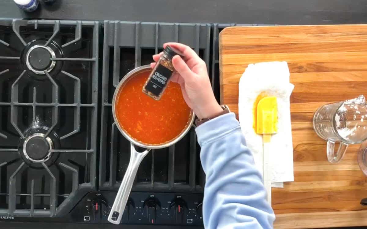 A person sprinkles pepper from a black bottle into a saucepan of red sauce on a stove. A wooden cutting board with a glass cup, a yellow spatula, and a paper towel is beside the stove.