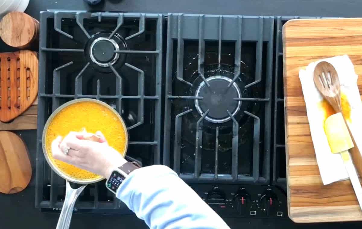 A person’s hand pours an orange liquid from a blender into a saucepan on a gas stove. A wooden spoon rests on a cutting board nearby. The stove has two burners, and small wooden trivets are visible on the left.