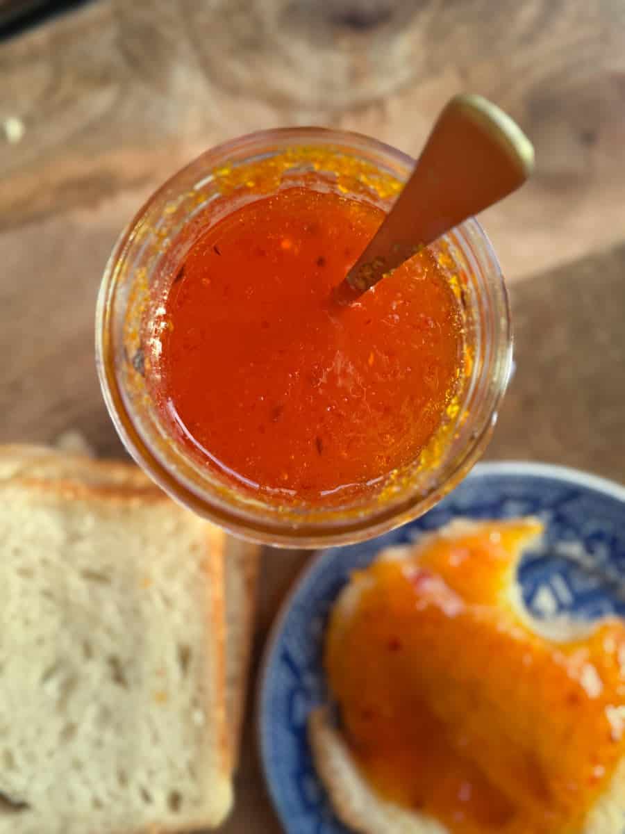 A jar of orange marmalade with a spoon inside is placed on a wooden surface. Next to it, there is a slice of bread and a blue-patterned plate with marmalade spread over another slice of bread.
