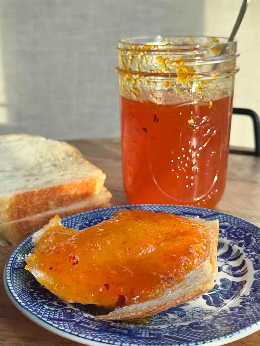 A slice of bread spread with orange marmalade sits on a blue and white plate. Behind it, a jar of marmalade with a spoon inside is partially full. Another slice of plain bread is adjacent to the jar, all set on a wooden surface.
