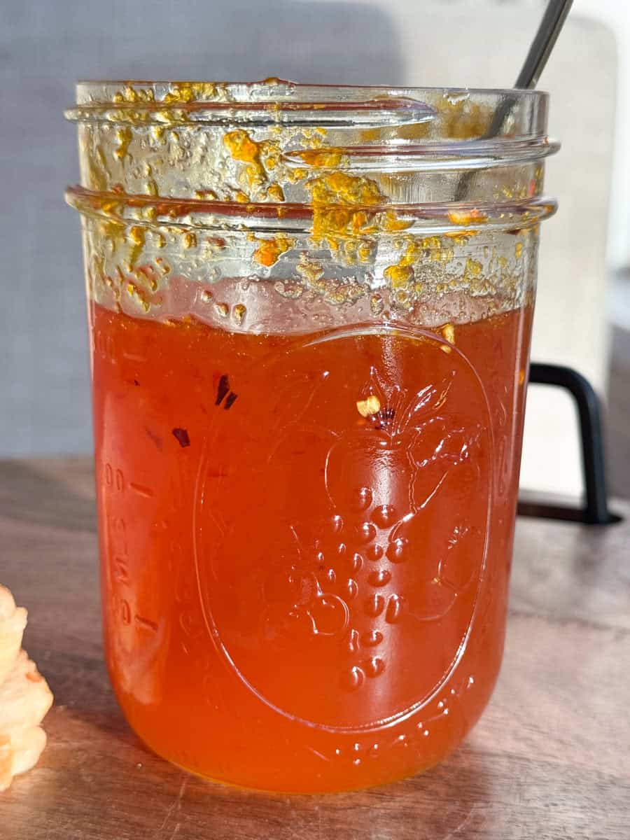 A glass jar filled with orange marmalade, showing remnants of spread on the upper inside. The jar features a raised fruit design. A metal spoon is inside. The jar is placed on a wooden surface, with sunlight casting shadows.