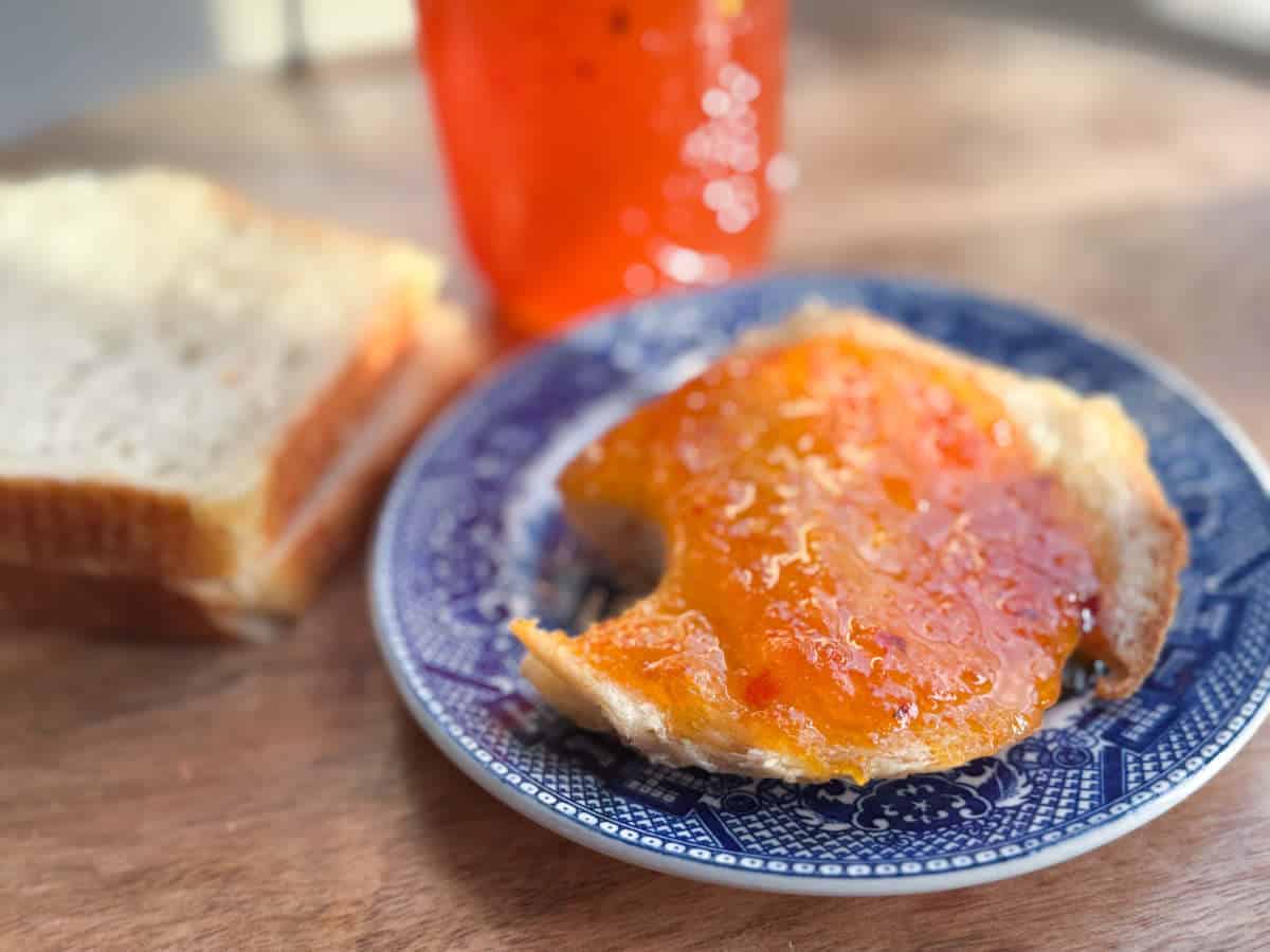 A close-up of a piece of toast spread with orange marmalade on a blue patterned plate. In the background, there is a slice of plain white bread and a jar of marmalade on a wooden surface.