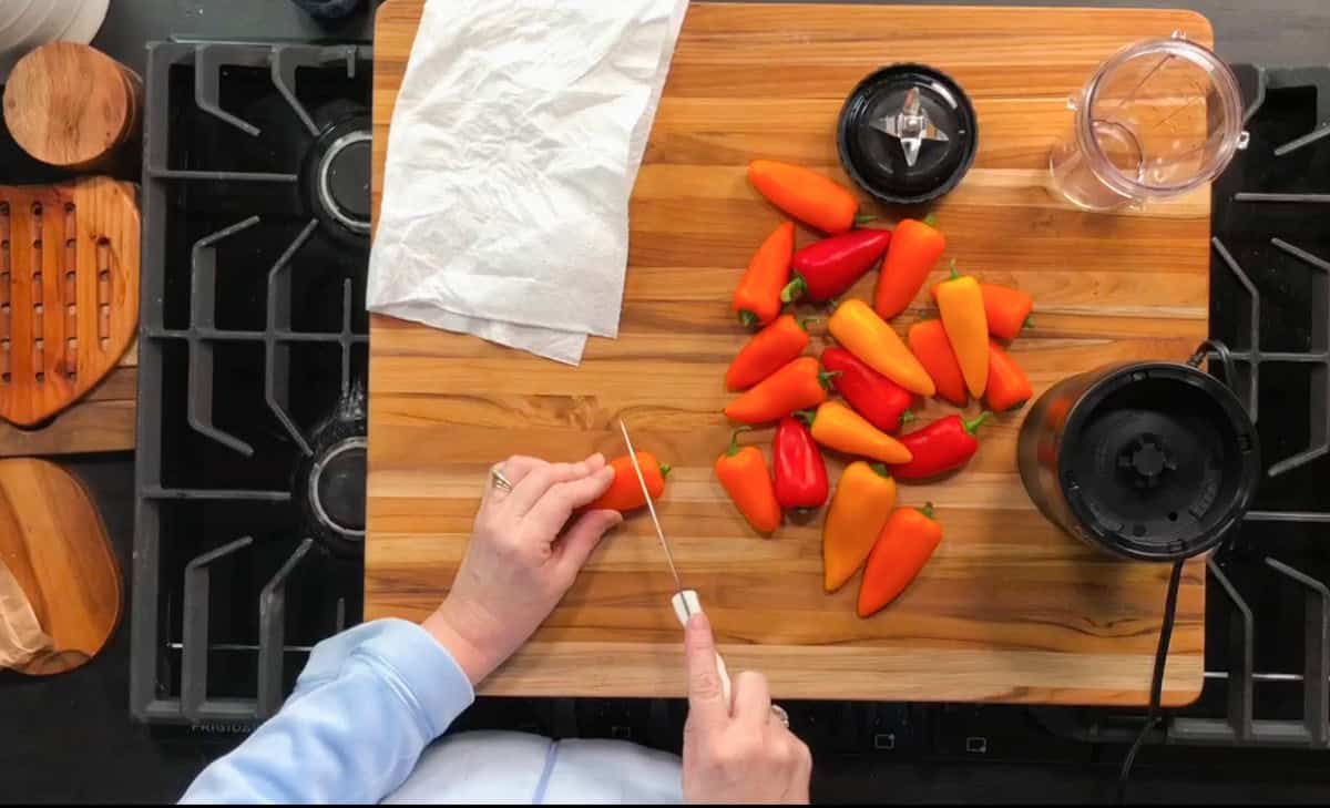 A person is slicing a small orange pepper on a wooden cutting board. There are red and orange peppers nearby. A blender base and cup, paper towel, and stove burners are also visible on the countertop.