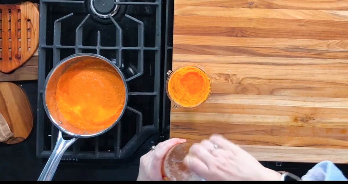 A person holds a glass jar beside a stove with a pot containing a bright orange liquid. Another jar, also with an orange liquid, is on a wooden countertop. Part of the stovetop is visible with a single burner in use.
