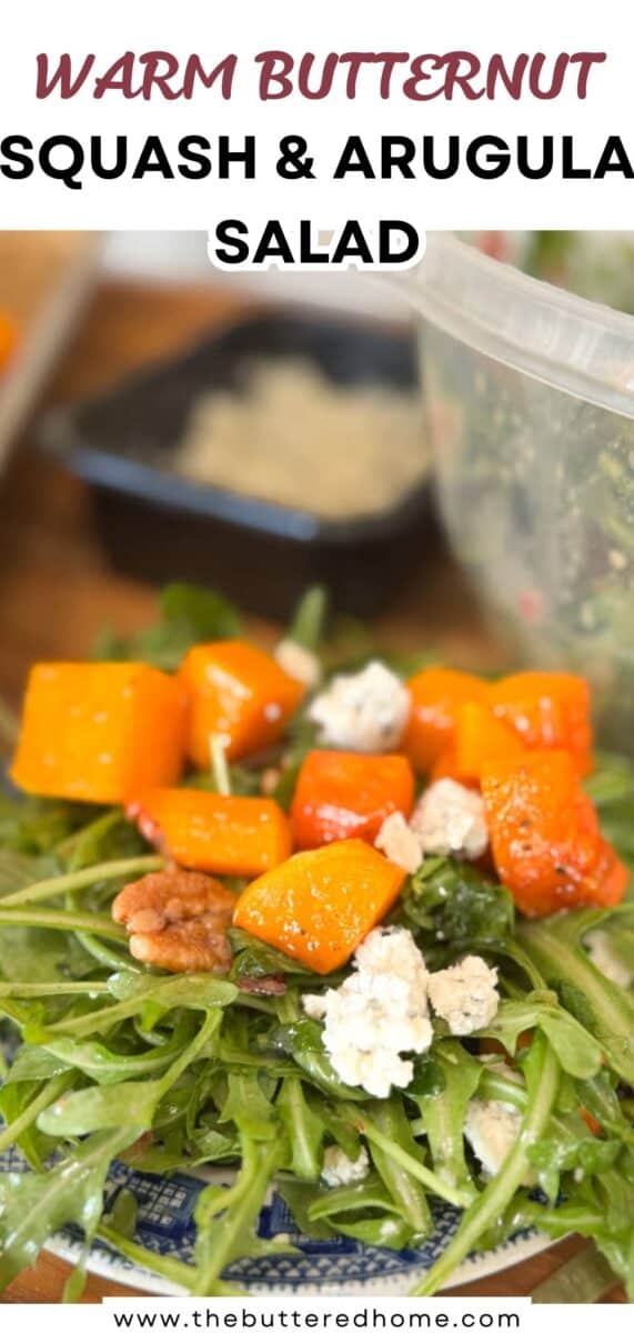A close-up of a salad featuring arugula, roasted butternut squash cubes, crumbled cheese, and walnuts on a wooden surface. A container of dressing is blurred in the background. Text above reads, "Warm Butternut Squash & Arugula Salad.