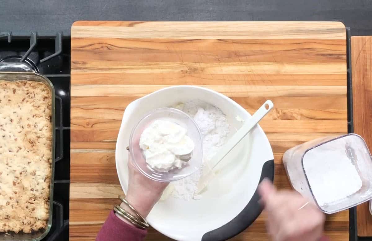 A person is adding a small plastic cup of a white substance, such as sour cream or yogurt, into a mixing bowl containing flour. A spatula rests in the bowl. A baked dish is on the stove to the left. A container of flour is to the right.