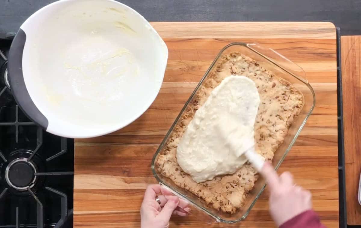 A person spreads cream cheese frosting over a rectangular baking dish filled with baked cookies or cake. The frosting is being smoothed with a spatula. A bowl with leftover batter sits on the wooden countertop beside a stovetop.