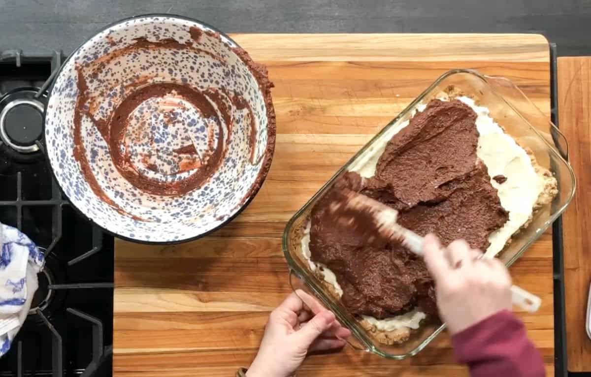 A person spreads chocolate batter over a creamy layer in a rectangular glass dish. Nearby, a speckled mixing bowl with remnants of the batter is on a wooden cutting board next to a stove.
