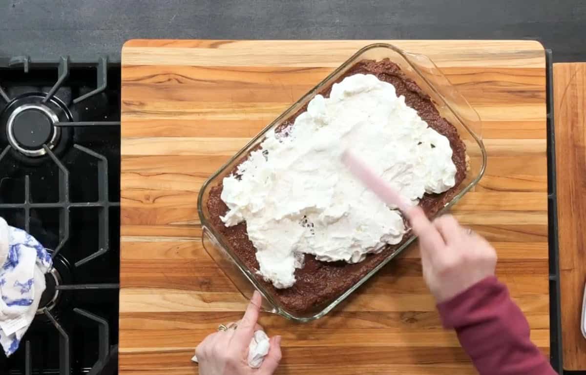 A person spreads whipped cream over a rectangular chocolate cake in a glass dish on a wooden cutting board. The board is next to a black stove with visible burner grates. A white and blue cloth is on the counter nearby.