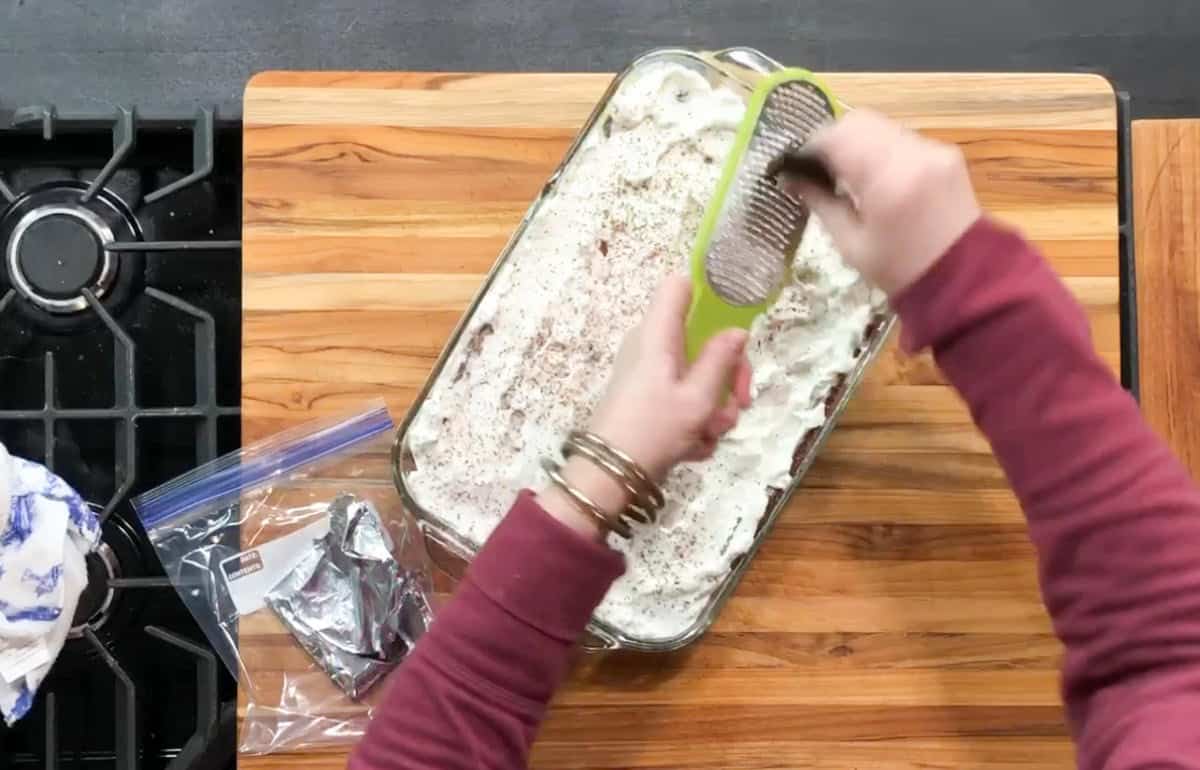 A person in a red sweater grates chocolate over a dish topped with white cream. The dish sits on a wooden cutting board next to a gas stovetop. A plastic bag with chocolate pieces and a cloth are nearby. The scene appears to be in a kitchen.