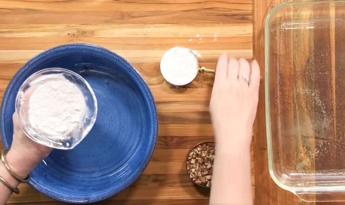 Hands pour flour from a small glass bowl into a larger blue bowl on a wooden surface. Nearby are a measuring cup with more flour and a small dish with chopped nuts. A clear baking dish is on the right.