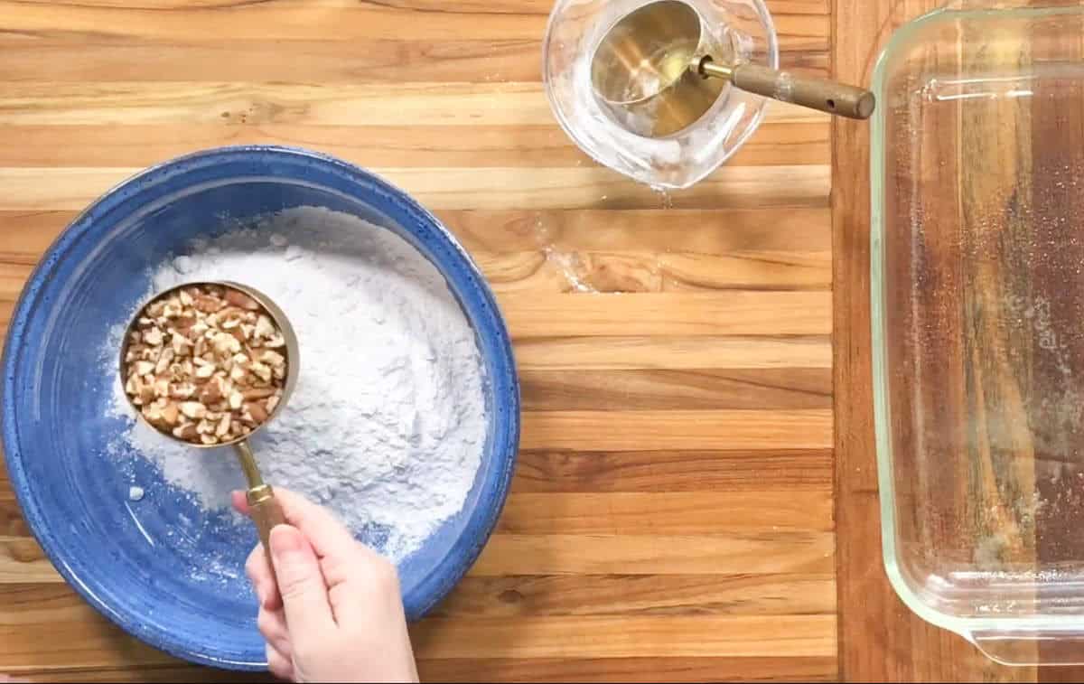 A hand holds a measuring cup with chopped nuts over a blue bowl containing flour. A glass dish and a small glass cup with water sit on a wooden countertop.