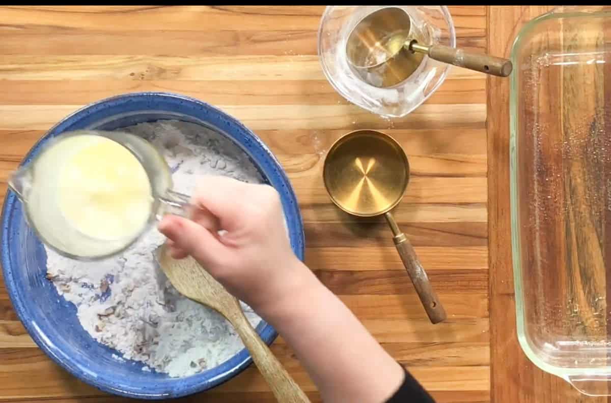 A person's hand pours melted butter from a glass measuring cup into a blue mixing bowl containing flour. A wooden spoon rests in the bowl. Nearby are a glass baking dish, a small glass bowl, and measuring cups on a wooden surface.