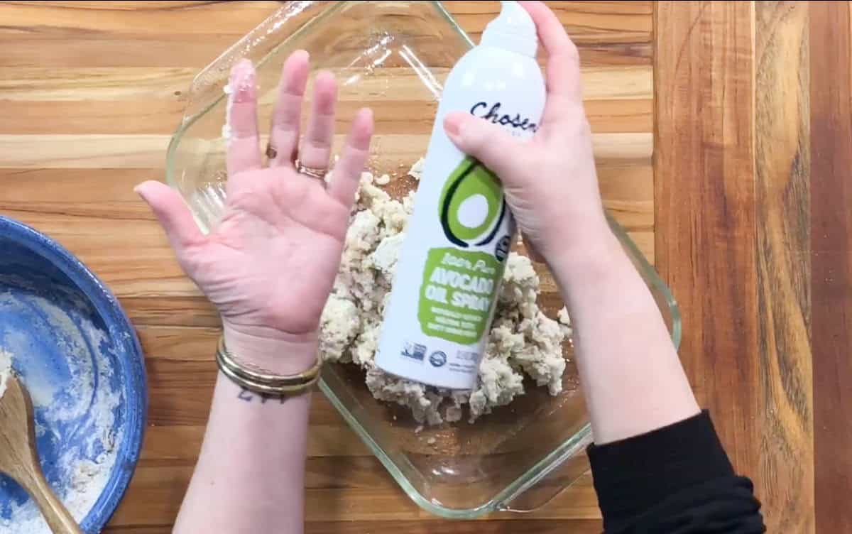 A person holds a can of avocado oil spray over a glass baking dish containing a crumbled mixture. A blue bowl with remnants of flour mixture is nearby, all set on a wooden counter. The person's wrist has multiple bracelets.