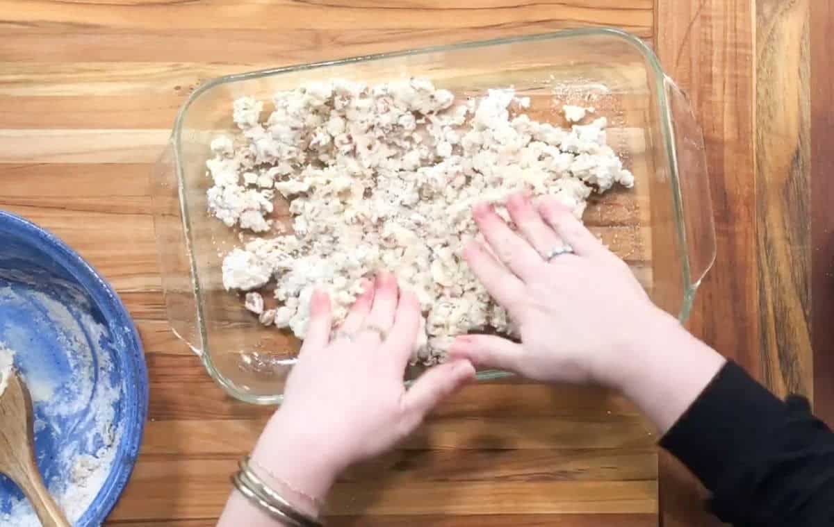 Hands are spreading a crumbly mixture in a glass baking dish on a wooden countertop. A blue bowl with a wooden spoon is on the left, containing some of the mixture. The person is wearing bracelets.