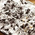 A close-up of a dessert topped with whipped cream and sprinkled with dark chocolate shavings. Crumbled bits of a light-colored ingredient are visible among the chocolate. The dessert is in a rectangular dish on a wooden surface.