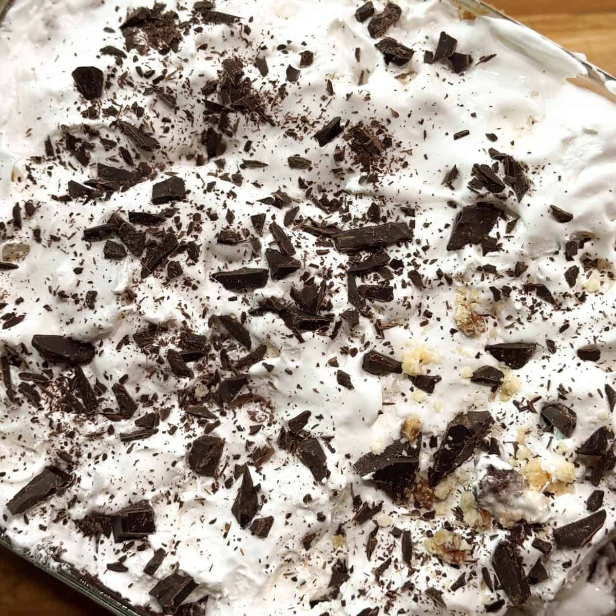 A close-up of a dessert topped with whipped cream and sprinkled with dark chocolate shavings. Crumbled bits of a light-colored ingredient are visible among the chocolate. The dessert is in a rectangular dish on a wooden surface.