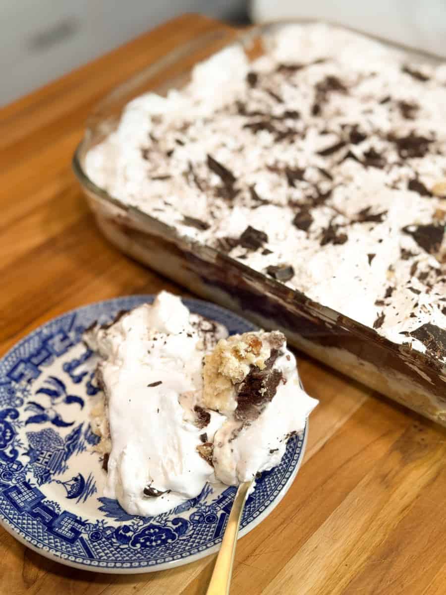 A dessert in a glass dish with layers of cream, cake, and chocolate shavings. A portion is served on a blue and white patterned plate, resting on a wooden surface. A gold-colored fork is placed on the plate.