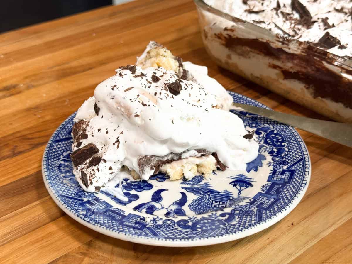 A piece of dessert on a blue and white plate. The dessert consists of layers with whipped topping and chocolate shavings. A larger portion in a glass dish with a serving utensil is in the background, all set on a wooden surface.