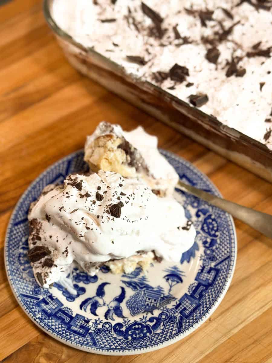A slice of dessert with whipped cream and chocolate shavings on a blue decorative plate sits next to a larger dish of the same dessert atop a wooden surface. A spoon rests beside the plate.