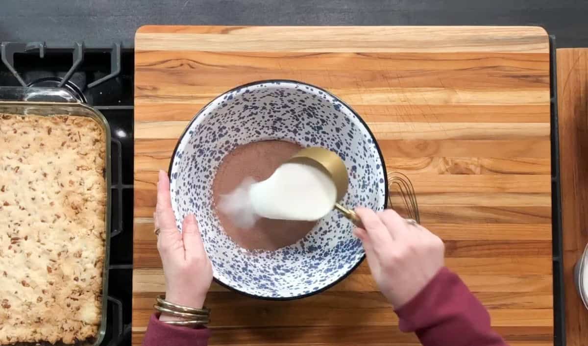 Person stirring ingredients in a large mixing bowl with a whisk. The bowl contains a pinkish powder and liquid being mixed. A baking dish with a cooked crust is on the left. The setting is a wooden countertop with a visible stovetop edge.