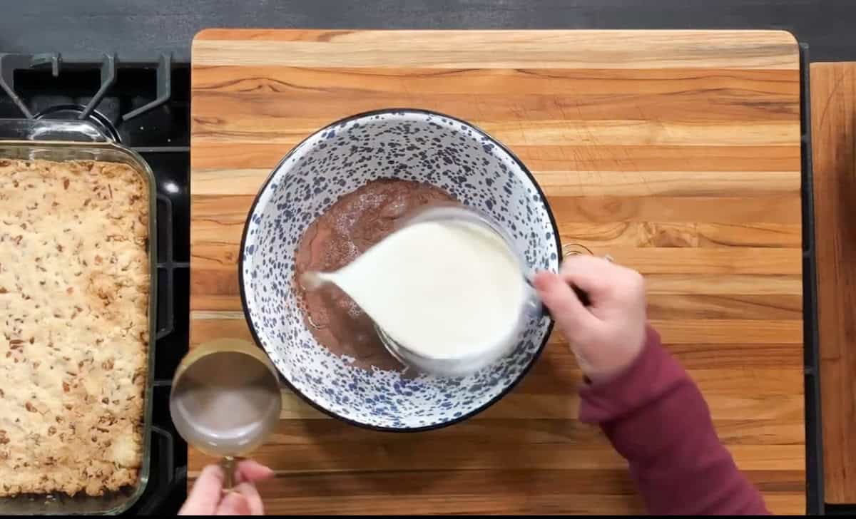 A person wearing a maroon sleeve pours milk from a measuring jug into a mixing bowl with chocolate batter. A measuring cup with remaining milk sits nearby. A baking dish with a crumbly dessert is on the left, all on a wooden countertop.