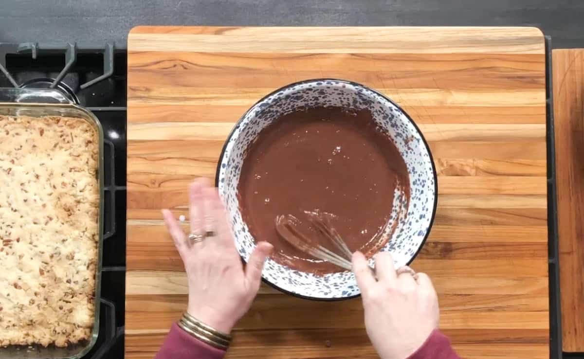 A person is whisking chocolate batter in a large white bowl with blue speckles on a wooden countertop. To the left, there is a glass dish with a baked crumbly dessert. The stove edge is visible on the left side.