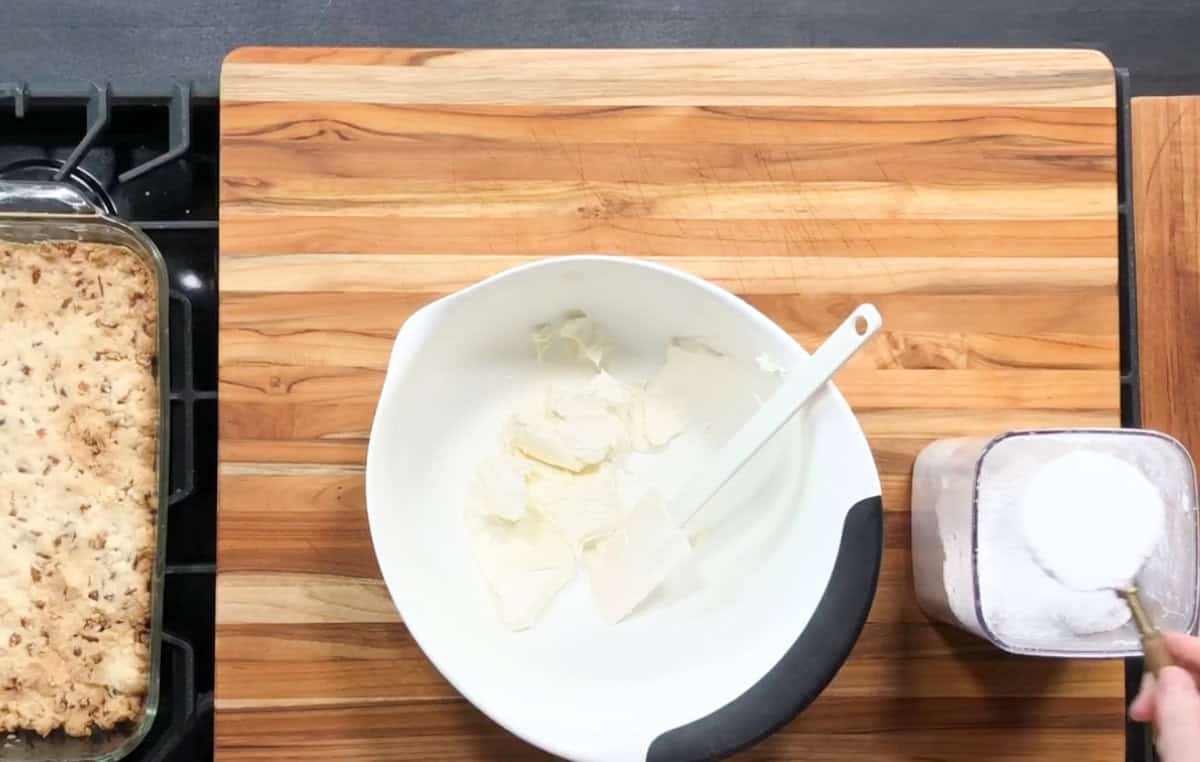 A wooden cutting board holds a white bowl with cream cheese and a spatula. Next to it, a hand is scooping sugar from a container. On the left, a baking dish filled with an oat-topped dessert rests on a stovetop.