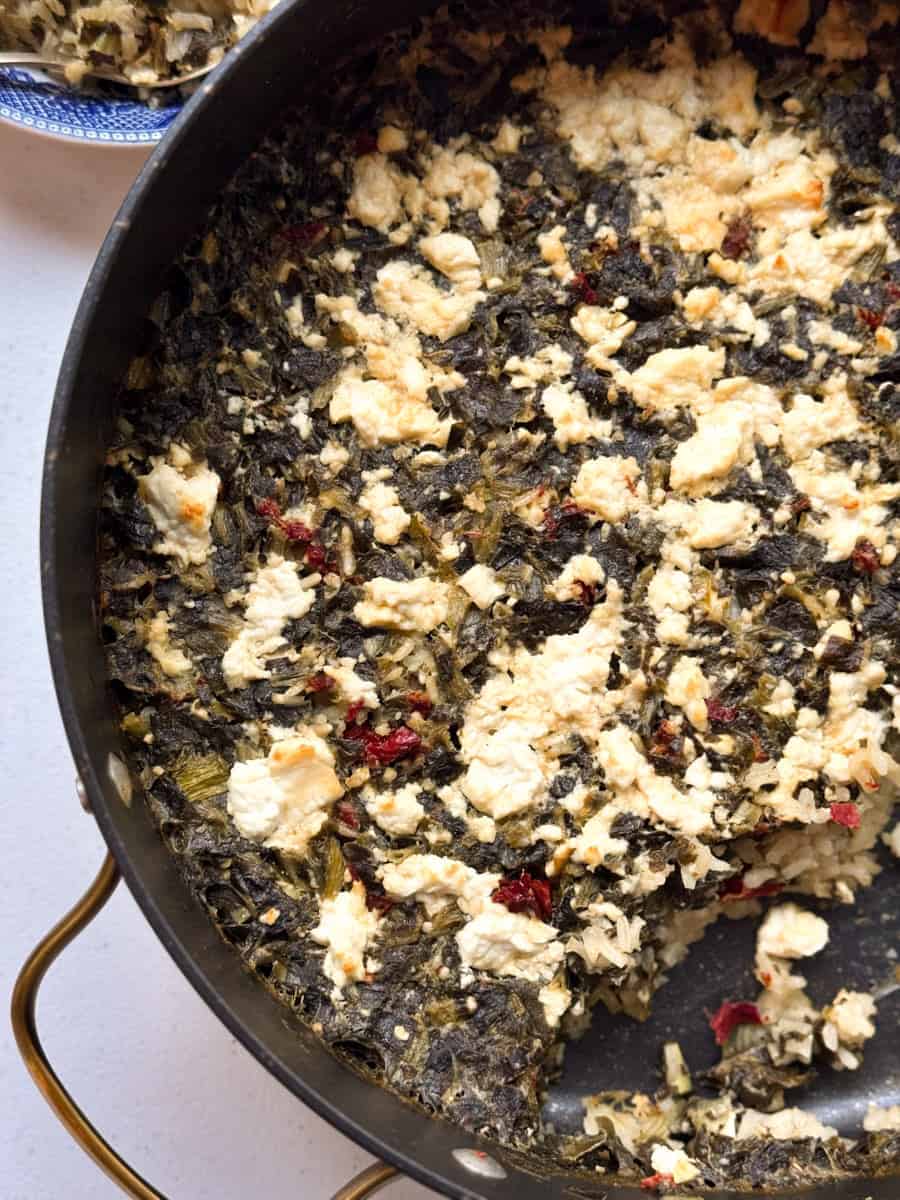 A round baking dish filled with a cooked mixture of leafy greens, crumbled cheese, and small red pieces, possibly vegetables or seasoning. A portion of the dish is missing, revealing the mixture's texture. A plate is partially visible in the background.