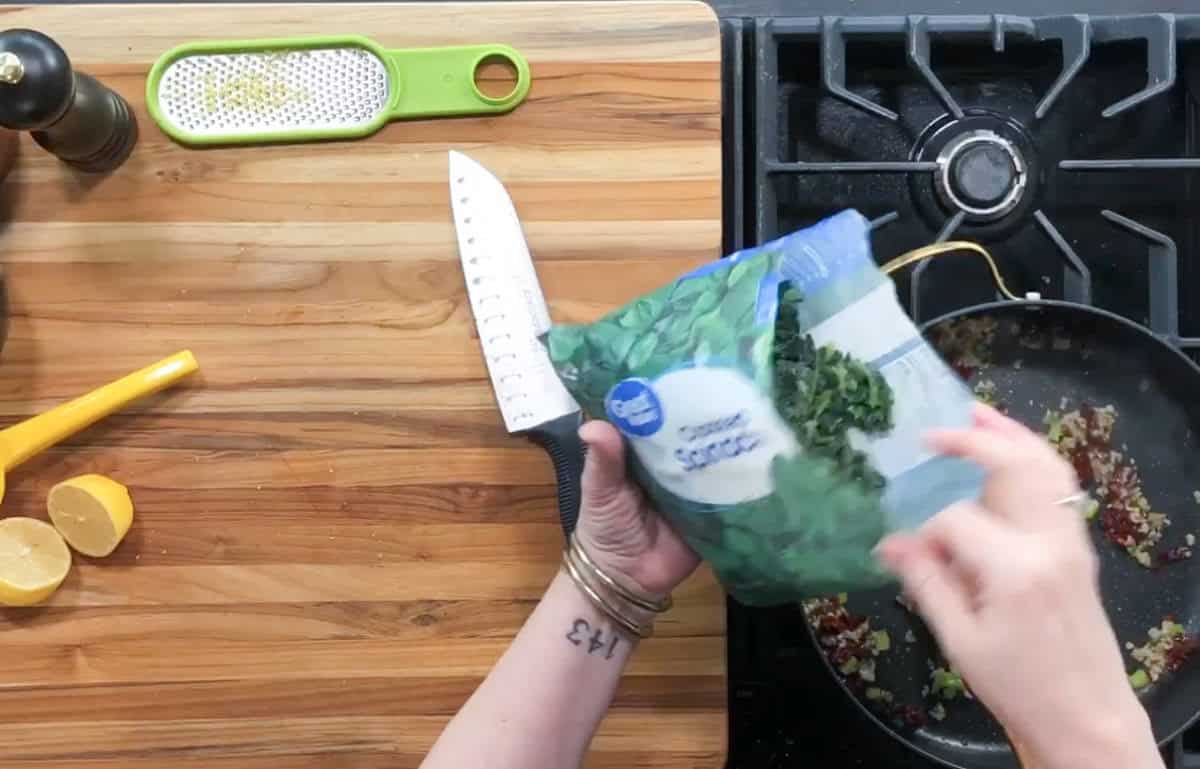 A person is adding chopped spinach from a blue bag into a pan on a black stove. A knife, a lemon, a grater, and a pepper grinder are on the wooden countertop. The person's wrist has a bracelet and a tattoo.