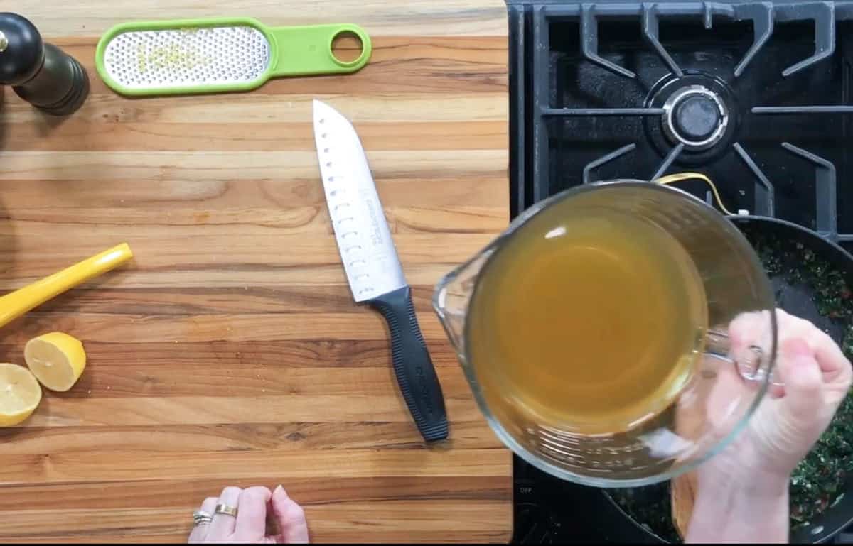 Overhead view of a kitchen counter with a cutting board holding a knife, lemon halves, and a grater. A person holds a measuring cup with a light brown liquid, pouring it into a pot on a stove. A pepper grinder is visible on the side.