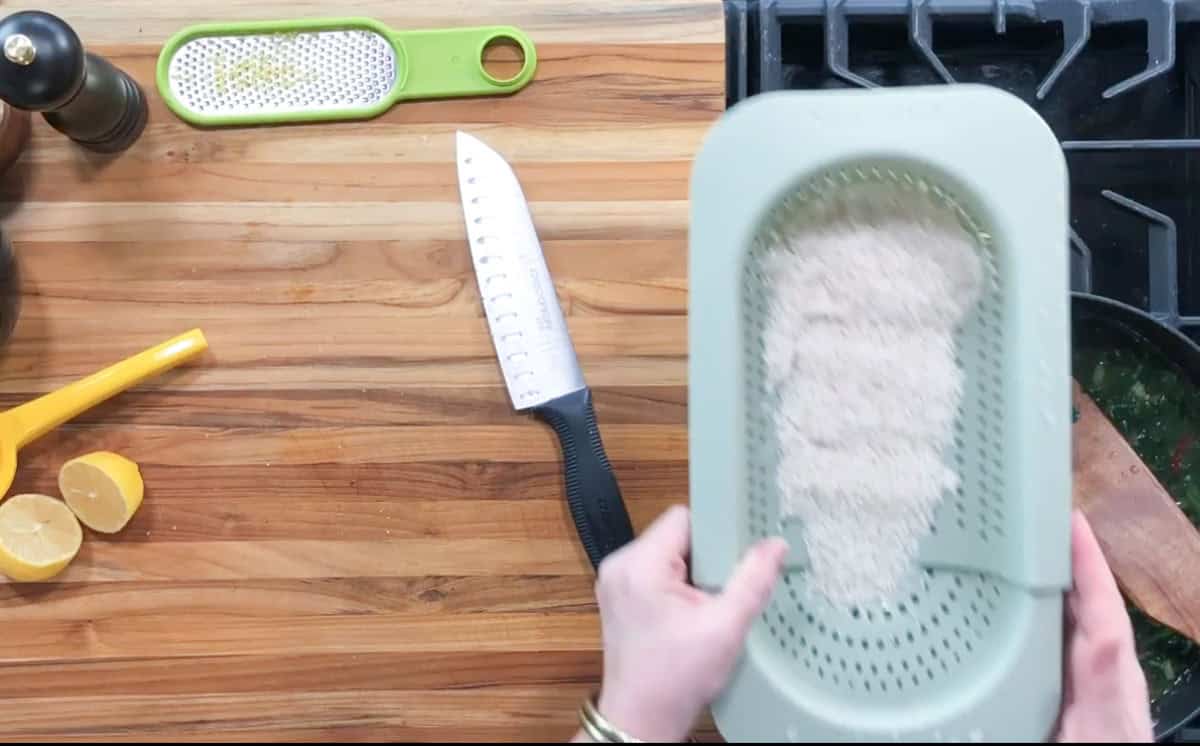 A person holds a green colander with rice over a wooden countertop. Nearby are a knife, a grater, lemon halves, and a yellow measuring cup. Part of a stovetop with a pan is visible on the right.