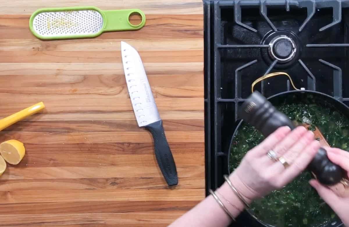A person is seen grinding pepper into a pan on a stove. To the left, a knife, lemon squeezer, lemon half, and grater rest on a wooden countertop. The pan contains a green, leafy dish. The scene suggests cooking in progress.