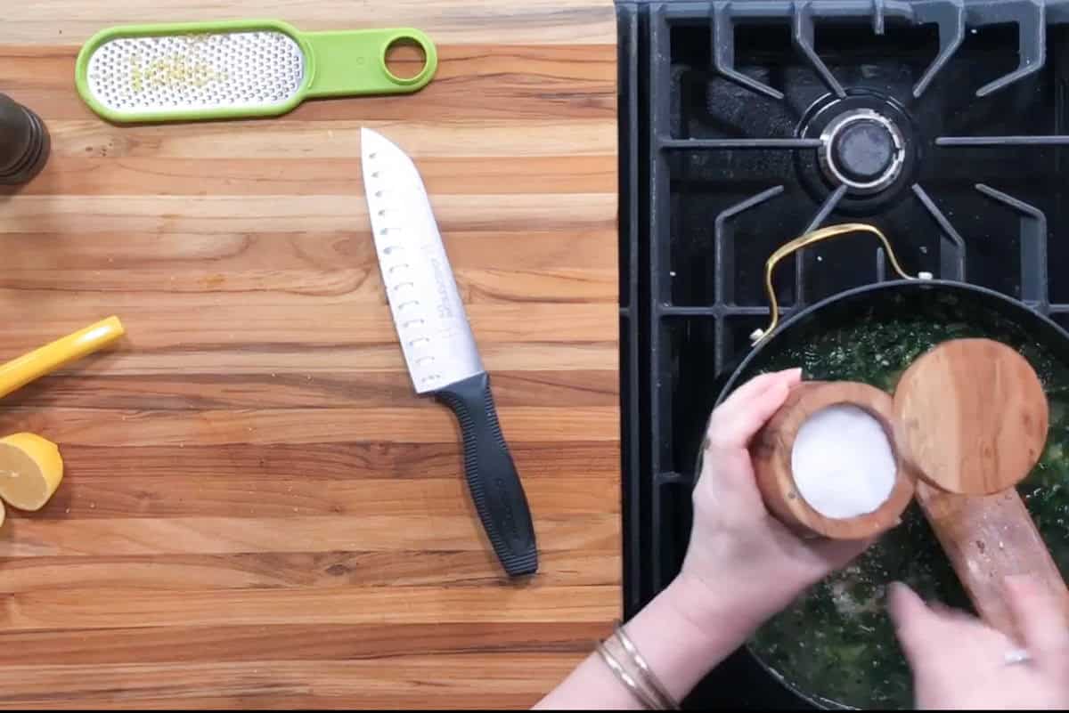 A person sprinkles salt from a wooden container into a pan on a stove. A cutting board with a knife, a grater, and a cut lemon are on the left. The pan on the stove contains chopped greens.