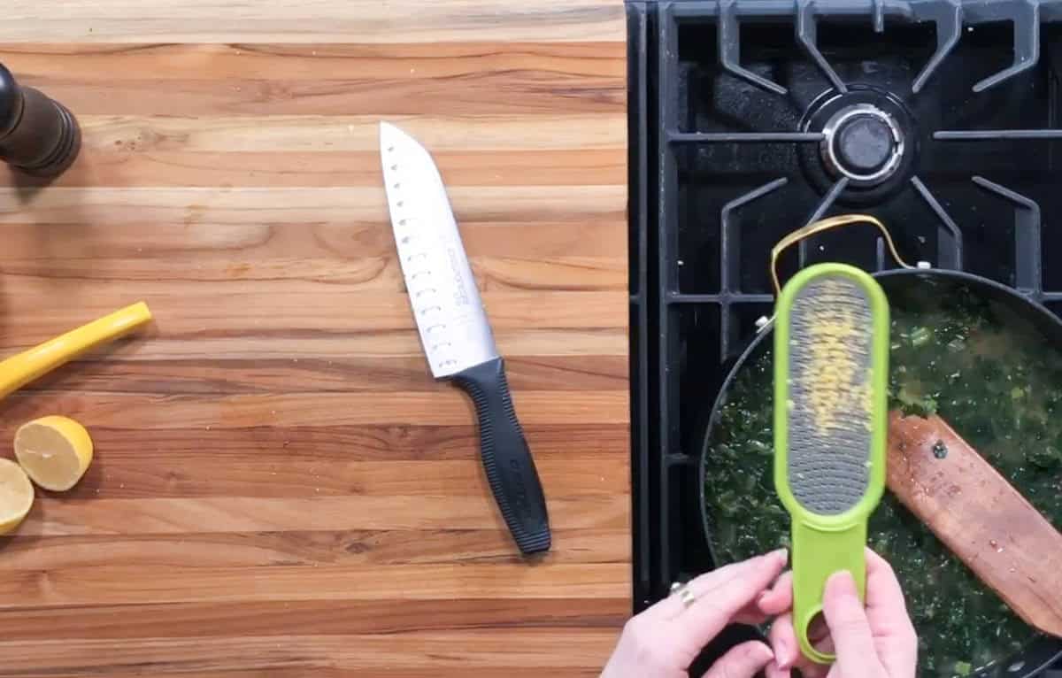 A person grates lemon zest over a pan with greens on a stove. A wooden spoon rests in the pan. A knife lies on a wooden countertop next to half a lemon and a yellow peeler.