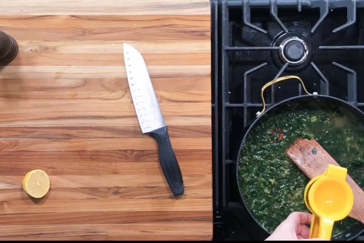 A kitchen scene showing a stovetop with a frying pan containing cooked greens and a piece of salmon. A hand is squeezing a lemon over the pan. A knife and half a lemon are on the wooden countertop nearby.