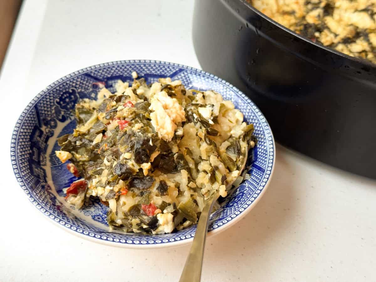 A blue and white bowl contains a dish of rice, cooked greens, and crumbled cheese. A spoon rests inside the bowl. The dish is placed on a white surface next to a black pot with more of the same food.