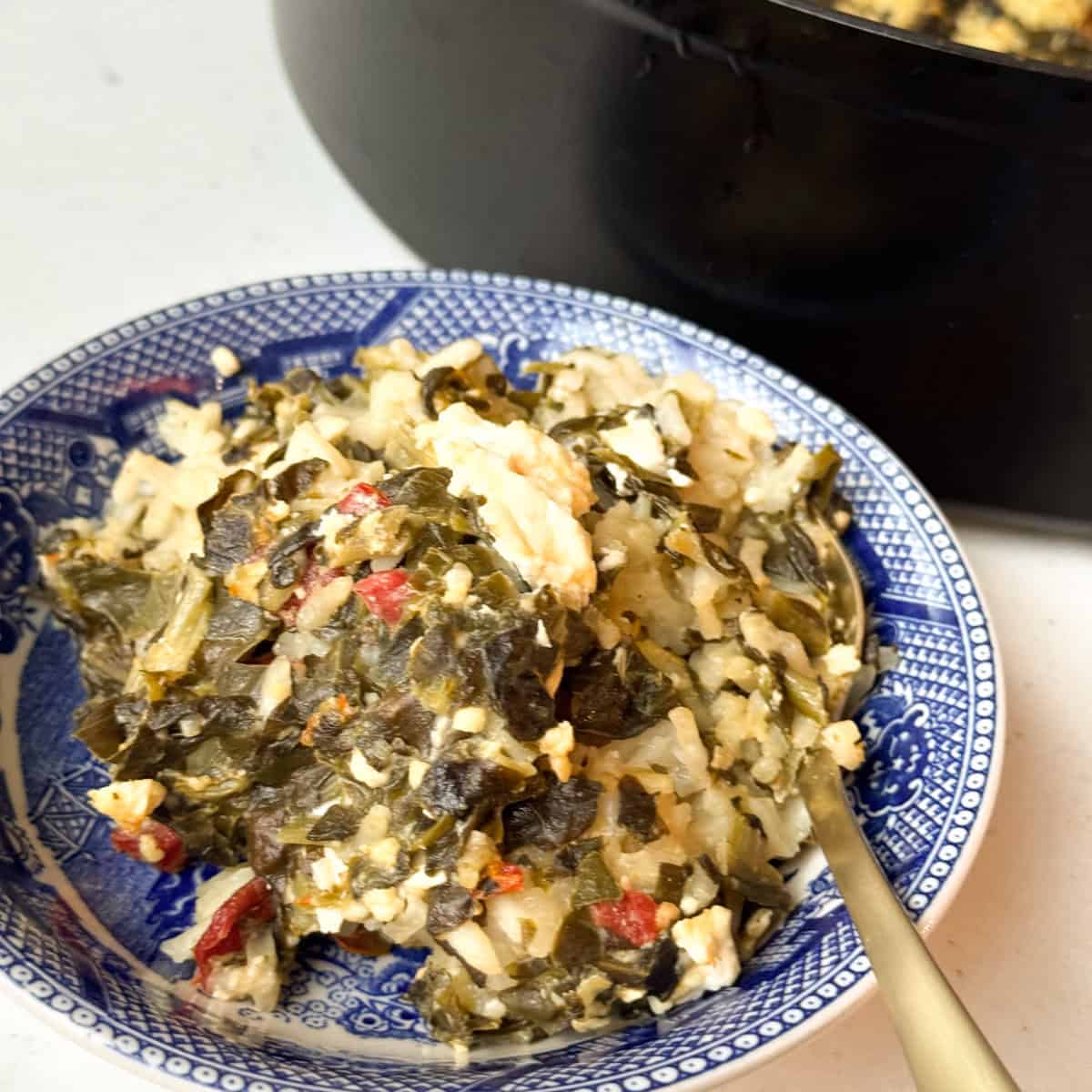 A serving of a rice and greens dish is on a blue-patterned plate, featuring chopped leafy greens, rice, red peppers, and crumbled cheese. A spoon is resting on the plate, and a black pot is partially visible in the background.
