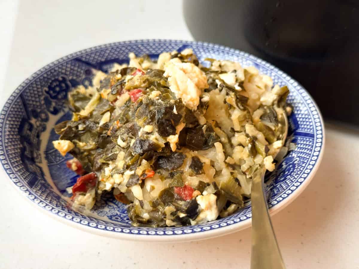A blue and white patterned bowl filled with a mixture of rice, cooked leafy greens, red peppers, and crumbled white cheese. A metal spoon rests on the side of the bowl. Part of a black container is visible in the background.