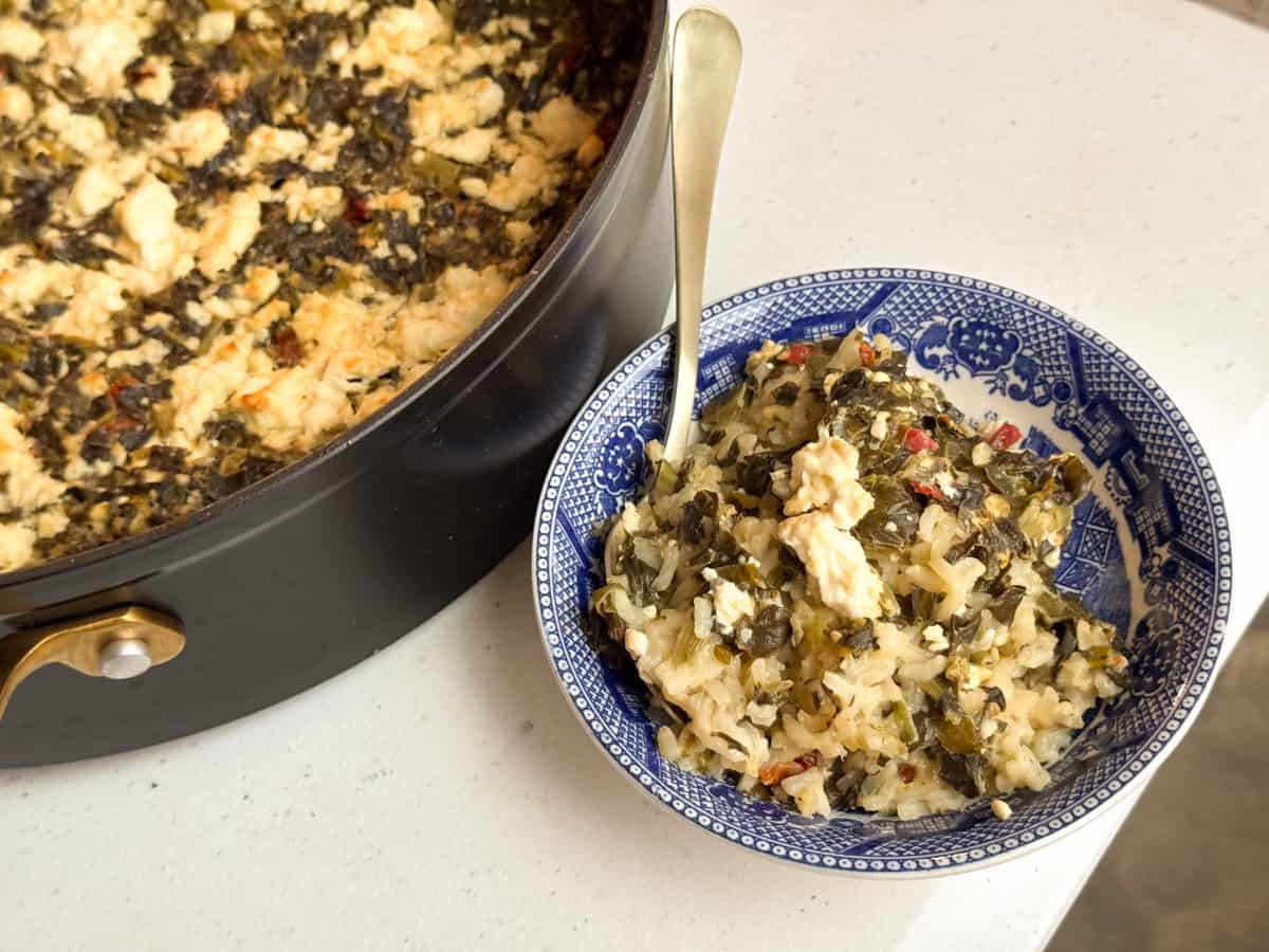 A close-up of a black pot and a blue-patterned bowl filled with a dish made of rice, spinach, and crumbled feta. A spoon is placed in the bowl. The background is a white countertop.