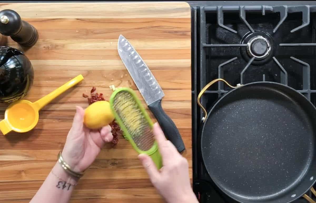A person is zesting a lemon with a green zester over a wooden cutting board. Nearby are chopped sun-dried tomatoes, a chef's knife, a yellow citrus juicer, a black pepper grinder, and an empty frying pan on a stove.