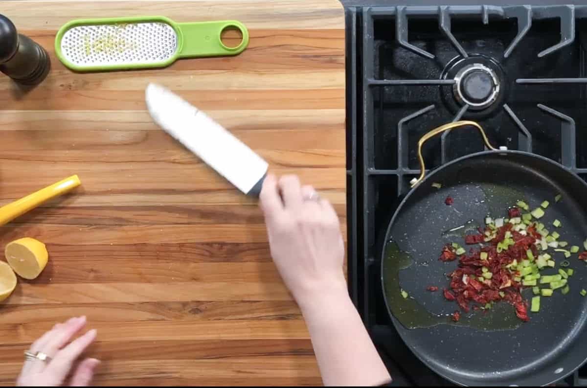 A person preparing food on a wooden countertop next to a black stove. A pan on the stove contains diced sun-dried tomatoes and green onions. Nearby are a grater, sliced lemon, and a knife held by the person, with a pepper mill to the left.
